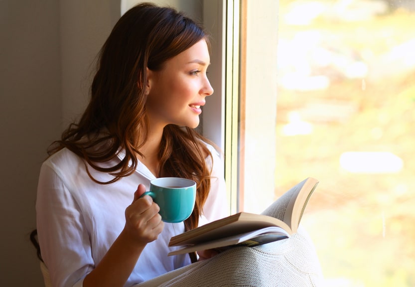 Donna rilassata che legge un libro alla finestra sorseggiando una tazza di té